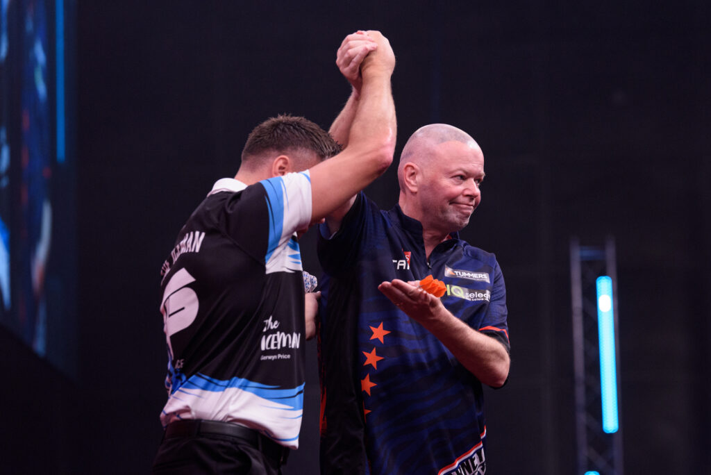Raymond van Barneveld hebt die Hand von Gerwyn Price in die Höhe, mit der freien Hand eine Präsentations-Geste.