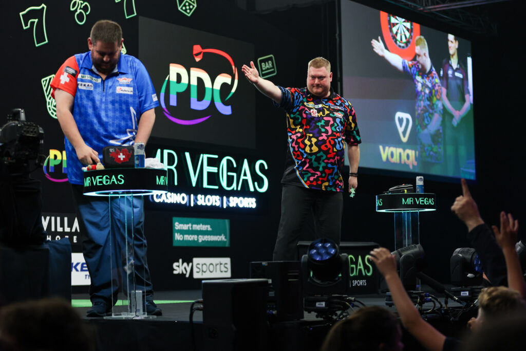 Stefan Bellmont am Tisch, im Hintergrund deutet Ricky Evans auf ihn.